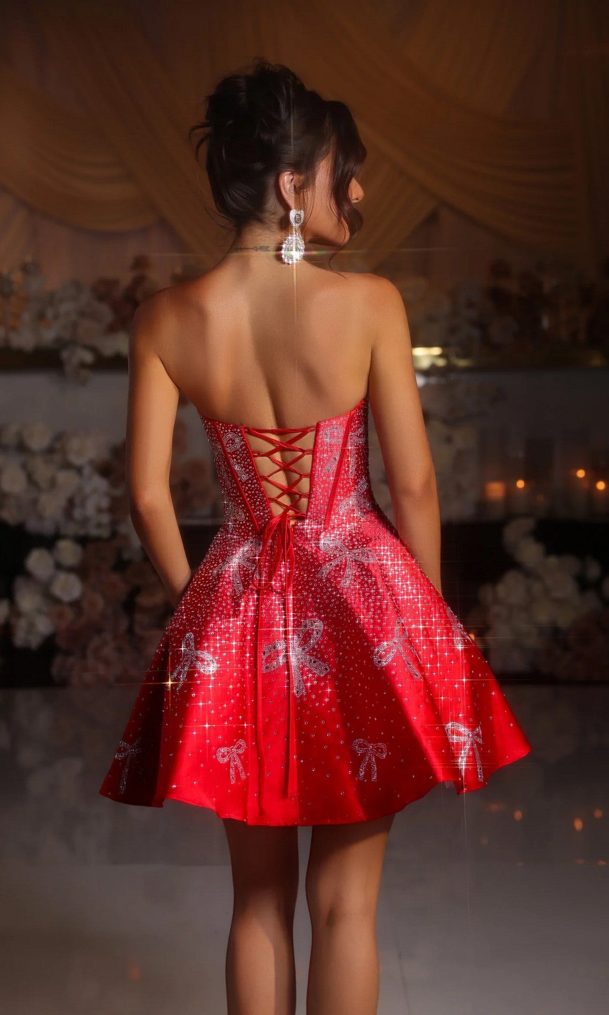 A woman stands indoors, facing away, wearing the Portia and Scarlett PS25785 short homecoming dress—a strapless fit-and-flare style with silver embellished bows and a lace-up back.